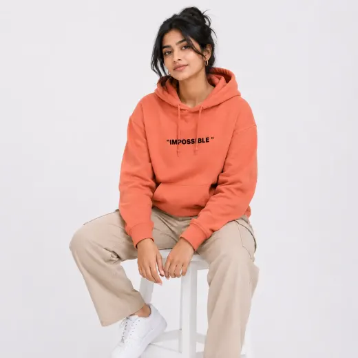Coral pullover hoodie with 'Never Say Die' print, hood and long sleeves, styled on a model in an urban setting